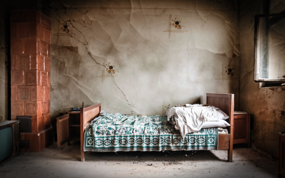 A twin size bed with blue printed coverlet and messy white pillows in a dingy old room with dented walls and old furniture.